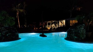 Une piscine illuminée la nuit, avec une personne allongée sur un flotteur, entourée de palmiers et d’un bâtiment dont les lumières se détachent sur le fond.
