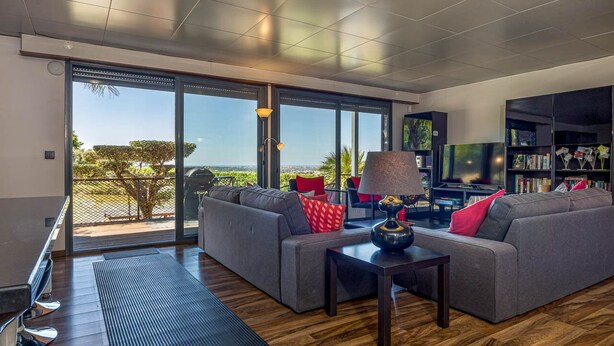 A modern living room with a grey sofa, a coffee table, and a TV. There is a bookshelf and a large window with a view of the outdoors. - Stunning 3 bedroom en-suite house in farm with pool (Estoi)