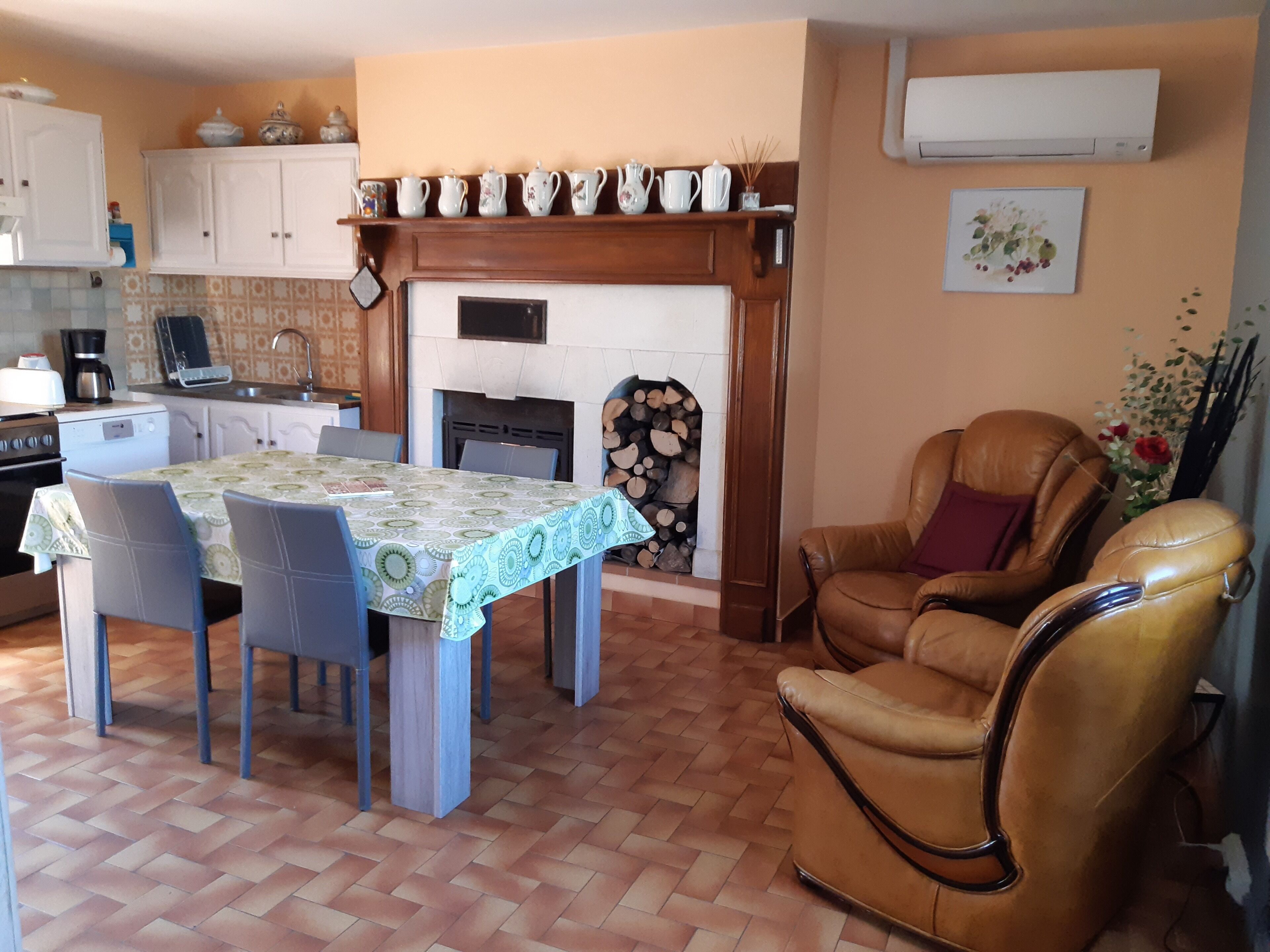 A cozy living room with a fireplace, a dining table with chairs, a leather armchair, and a kitchen area with cabinets and a coffee maker.
