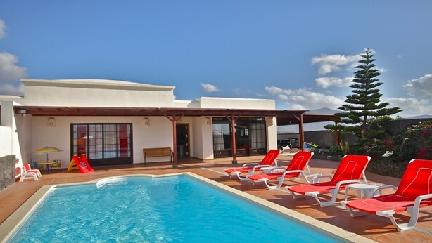 Ein Poolbereich mit klarem, blauem Pool, roten Liegestühlen und einem Gebäude mit großen Fenstern und einer überdachten Terrasse.