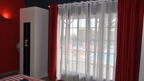 Ein Hotelzimmer mit Bett, Schrank, Fenster mit leichtem Vorhang und Blick auf einen Poolbereich.