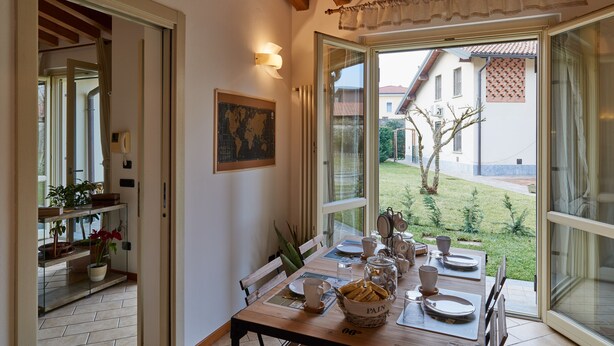 A dining area with a wooden table set for four, a window with a view of a garden and buildings, and a framed picture on the wall. - bcube - bed and breakfast in the village (Pavia)