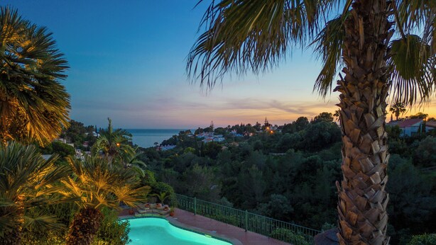 Ein Pool mit geschwungener Kante, umgeben von Palmen und Blick auf das Meer im Abenddämmerung.