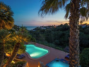 A swimming pool with a curved edge, surrounded by palm trees and a view of the sea at dusk. - 5 Bedroom Villa With Pool, Hot Tub & Stunning Sea View. Close to town and beach (Carvoeiro)