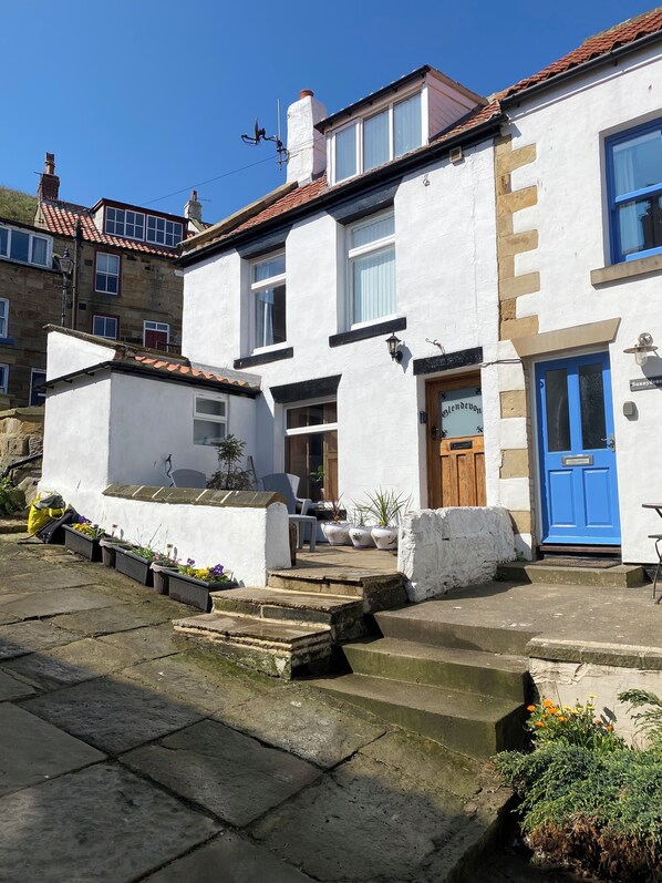 A white building with a blue door, a small garden area with potted plants, and a clear blue sky.