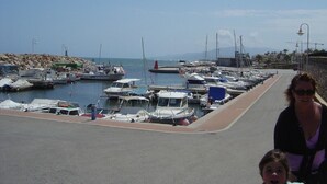 Uma marina com vários barcos atracados, um calçadão pavimentado e um litoral rochoso ao fundo.