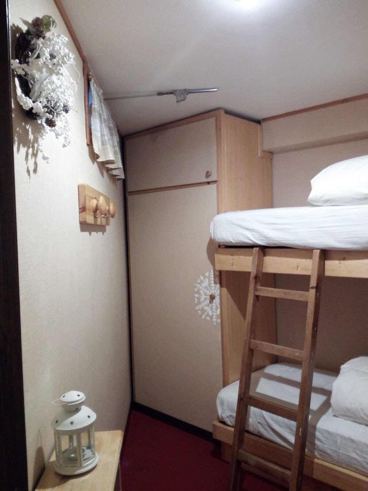 Una habitación pequeña y compacta con una cama litera, una escalera de madera, una mesita con un farolillo y un estante en la pared con adornos.