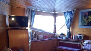 Een gezellig blokhutinterieur met een houten plafond, een rode bank, een houten bankje, een televisie op een kast, en een raam met geruit gordijnen.