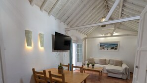 A spacious room with a high ceiling, a dining table with chairs, a sofa, a television mounted on the wall, and a door leading outside.