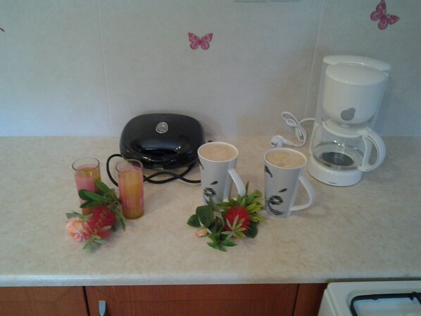 A kitchen counter with a coffee maker, two mugs, a glass of juice, and decorative flowers.