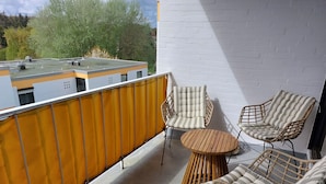 Ein Balkon mit Korbstühlen und einem runden Holztisch, eine weiße Backsteinmauer sowie Blick auf Bäume und Gebäude.