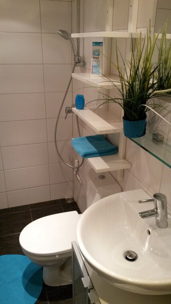 Une salle de bain équipée de toilettes, d’un lavabo, d’une douche et d’une étagère sur laquelle se trouvent une plante et un flacon de shampoing.