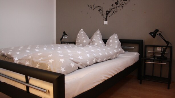 A neatly made bed with star-patterned bedding, two bedside tables with lamps, and a wall decoration. - Cozy little apartment (Südwest)