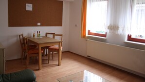 A room with a wooden dining table, chairs, a glass coffee table, a corkboard on the wall, and a window with curtains. - Cozy little apartment (Südwest)