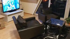 A modern living room with a television, a sofa, a coffee table, and a view of a fire through a glass door.