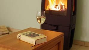 Una estufa de leña con puerta de cristal, una copa de vino blanco y un libro sobre una mesa de madera.