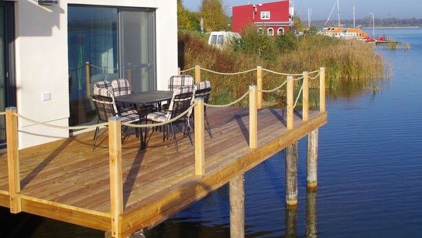 Un edificio moderno con una terraza de madera que se extiende sobre el agua, con un comedor que incluye sillas y una mesa.