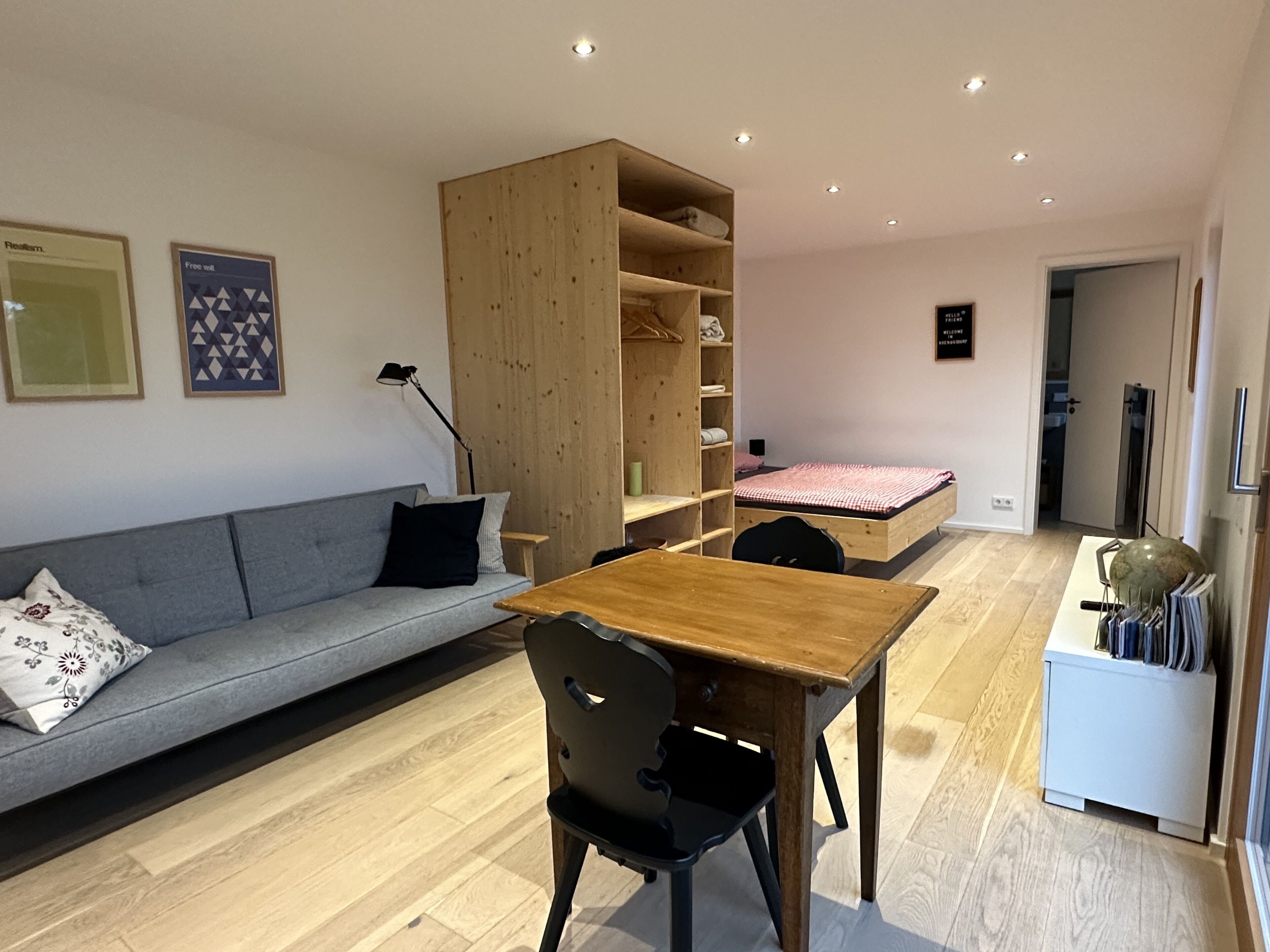 Un espacio habitable moderno y compacto con sofá de madera, una pequeña mesa de comedor, estantería empotrada y una cama individual.