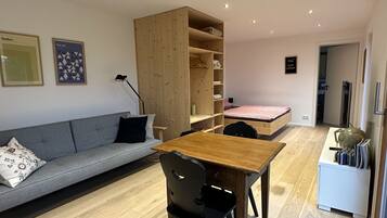 Un espacio habitable moderno y compacto con sofá de madera, una pequeña mesa de comedor, estantería empotrada y una cama individual.