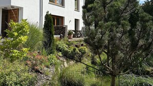 Een huis met twee verdiepingen, met een witte gevel, een balkon en een verzorgde tuin met verschillende planten en een kleine boom.