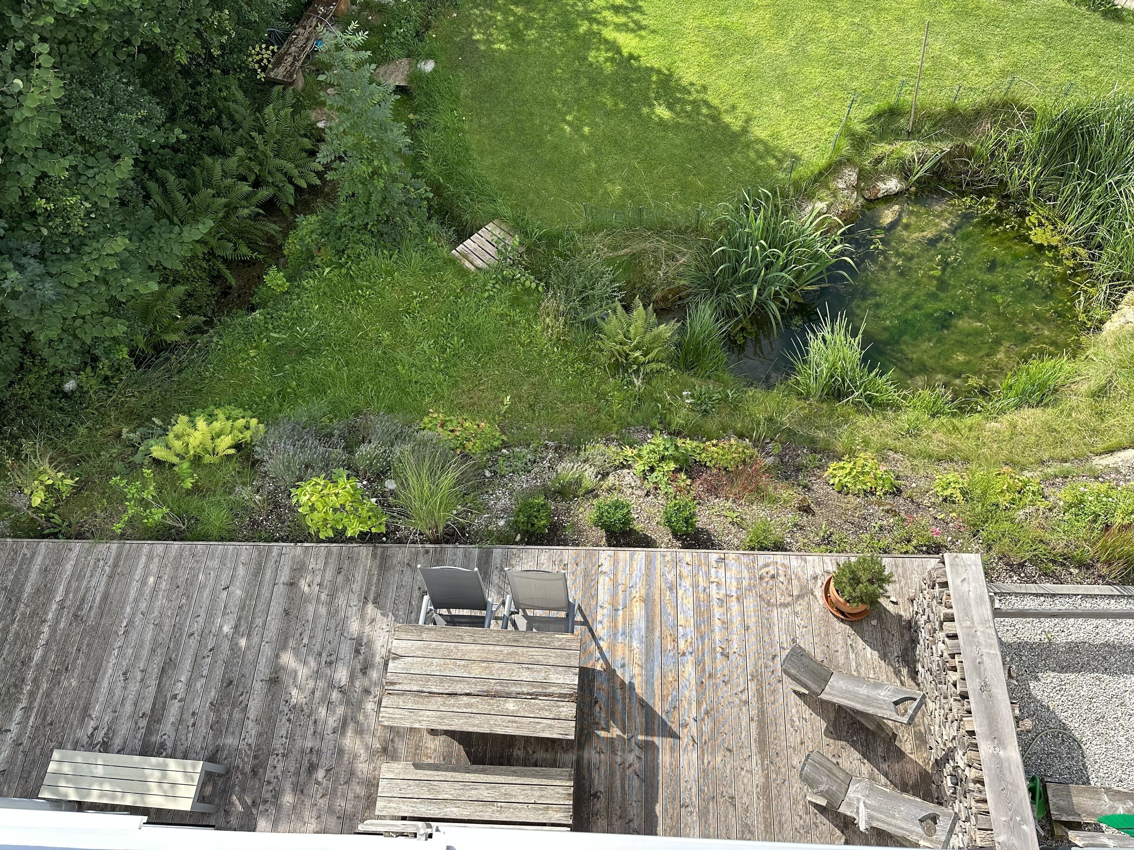 A wooden deck with a table and chairs, surrounded by a lush garden with various plants and a small pond.