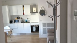 Moderni keittiö, jossa on valkoiset kaapit, mikroaaltouuni ja liesi. Keittiössä on myös ruokailutila, jossa on pöytä ja tuolit. Seinällä näkyy naulakko.