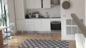 Moderni keittiö, jossa on valkoiset kaapit, sisäänrakennettu uuni ja mikroaaltouuni. Lattialla on kuvioitu matto ja pöydällä vihreä kansio.