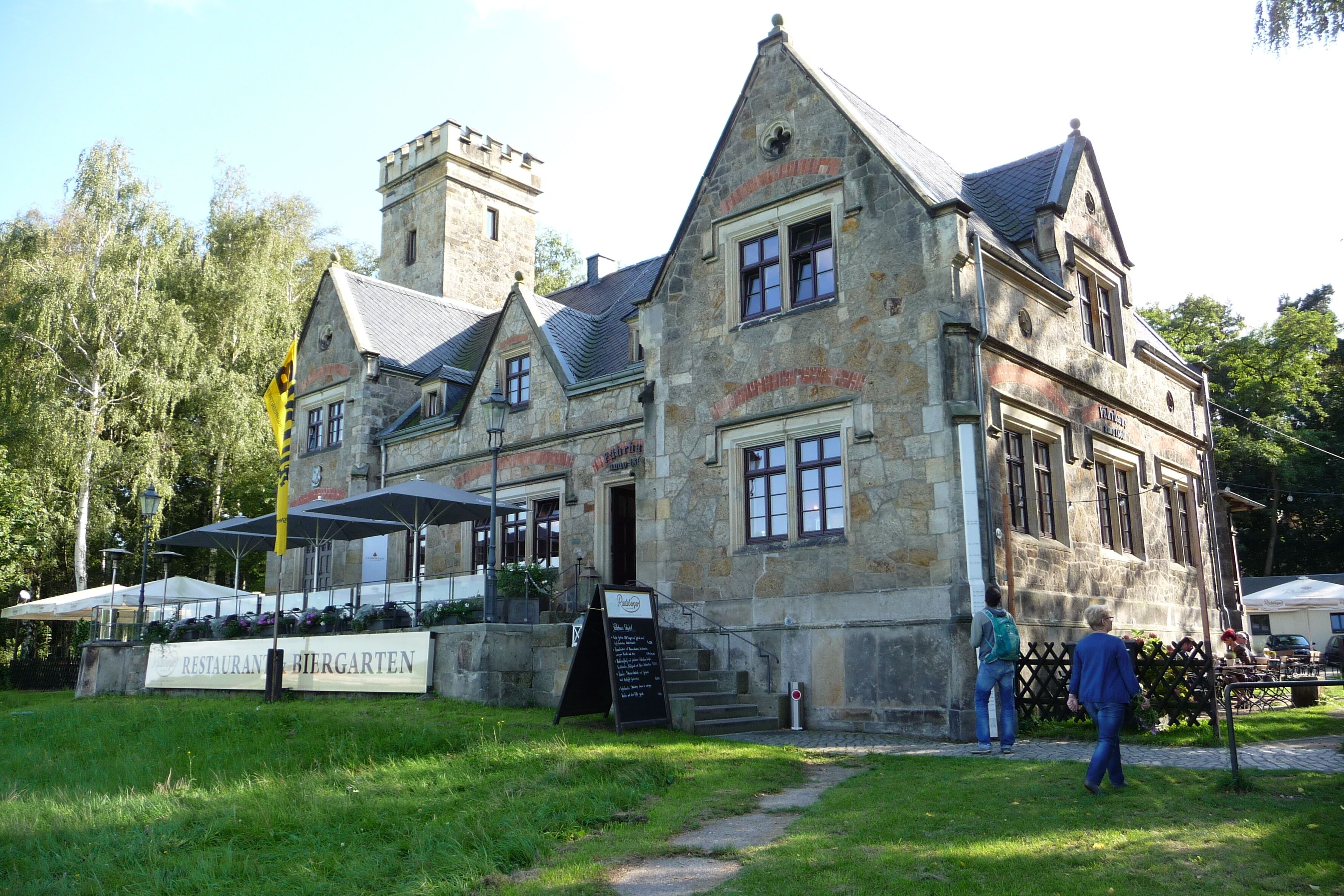 Ein historisches Steinhaus mit einem Turm, einem Schild mit der Aufschrift 'Restaurant Biergarten' und Sitzgelegenheiten im Freien mit Sonnenschirmen.