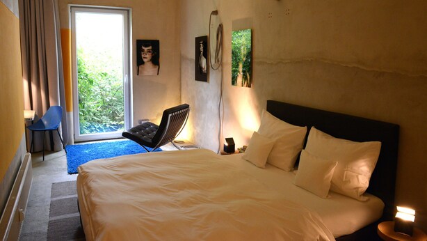 A hotel room with a large bed, a desk with a chair, a window with curtains, and artwork on the walls.