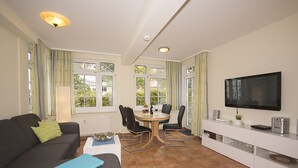 A modern living room with a sofa, a dining table with chairs, a TV mounted on the wall, and large windows with curtains.