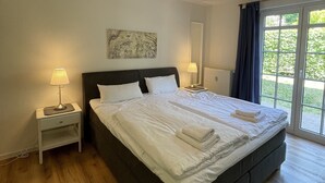 A large bed with white linens, a bedside table with a lamp, a ceiling light fixture, and a view of greenery through a window.