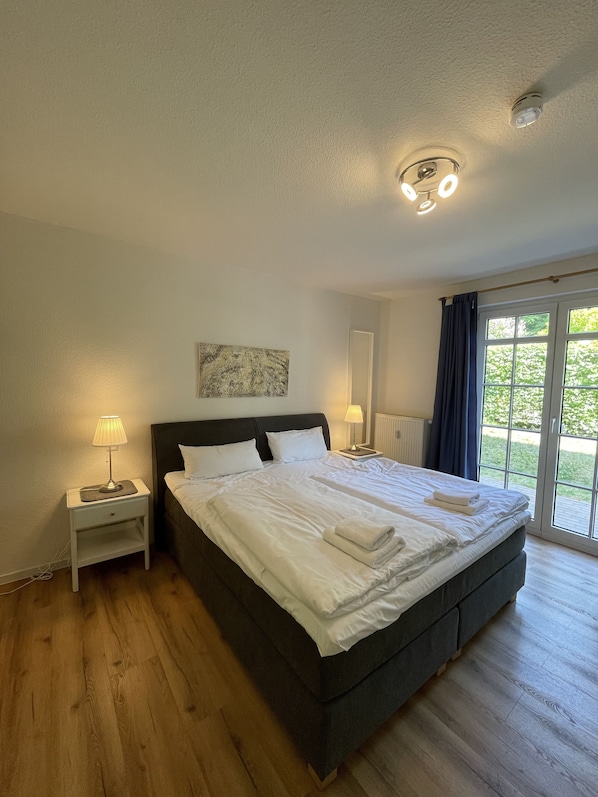 A large bed with white linens, a bedside table with a lamp, a ceiling light fixture, and a view of greenery through a window.