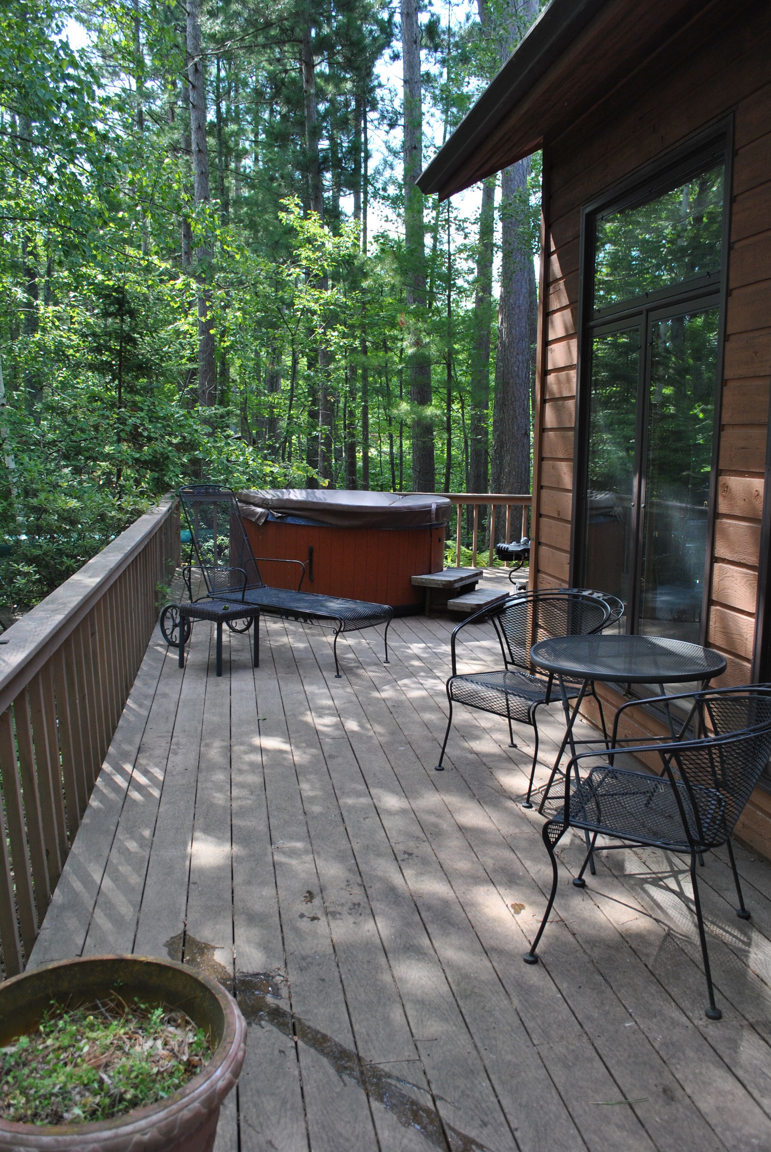 Une terrasse en bois, agrémentée de chaises et d’une table en métal, entourée d’une balustrade en bois, et offrant une vue sur une zone boisée.