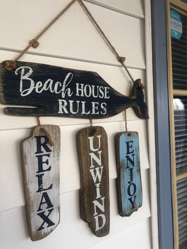 A wooden sign with 'Beach House Rules' and three smaller signs below it reading 'RELAX,' 'UNWIND,' and 'ENJOY.'
