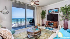 A living room with a large window overlooking the beach, a ceiling fan, a TV, and a small table with books and a picture frame. - Spectacular -  Beach front condo from 149 night - no extra administrative fees. (Panama City Beach)