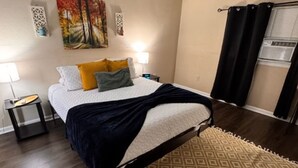 Une chambre à coucher avec un lit, une table de chevet, une lampe, un tableau au mur et une fenêtre avec des rideaux.