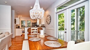 A dining area with a wooden table, woven placemats, white chairs, a chandelier, and a view of the outdoors through large windows. - Live oak views from this 3 bedroom cottage; 2 golf carts and peaceful screened in porch (Bald Head Island)