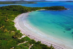An aerial view of a beach with clear turquoise waters, a sandy shoreline, and lush greenery.