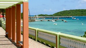 A coastal walkway with a green railing, a pier in the background, and boats on the water.
