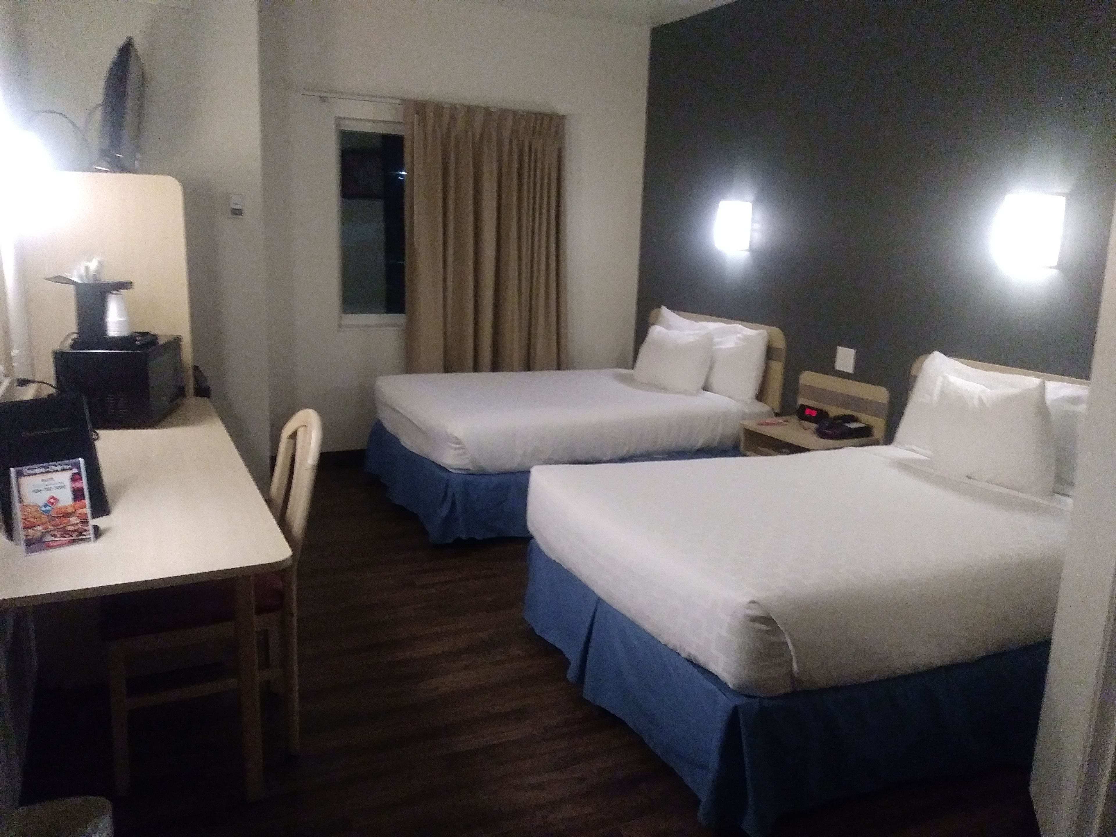 Une chambre d’hôtel avec deux lits simples, un bureau avec une chaise, une petite table, un téléviseur et une fenêtre avec des rideaux.