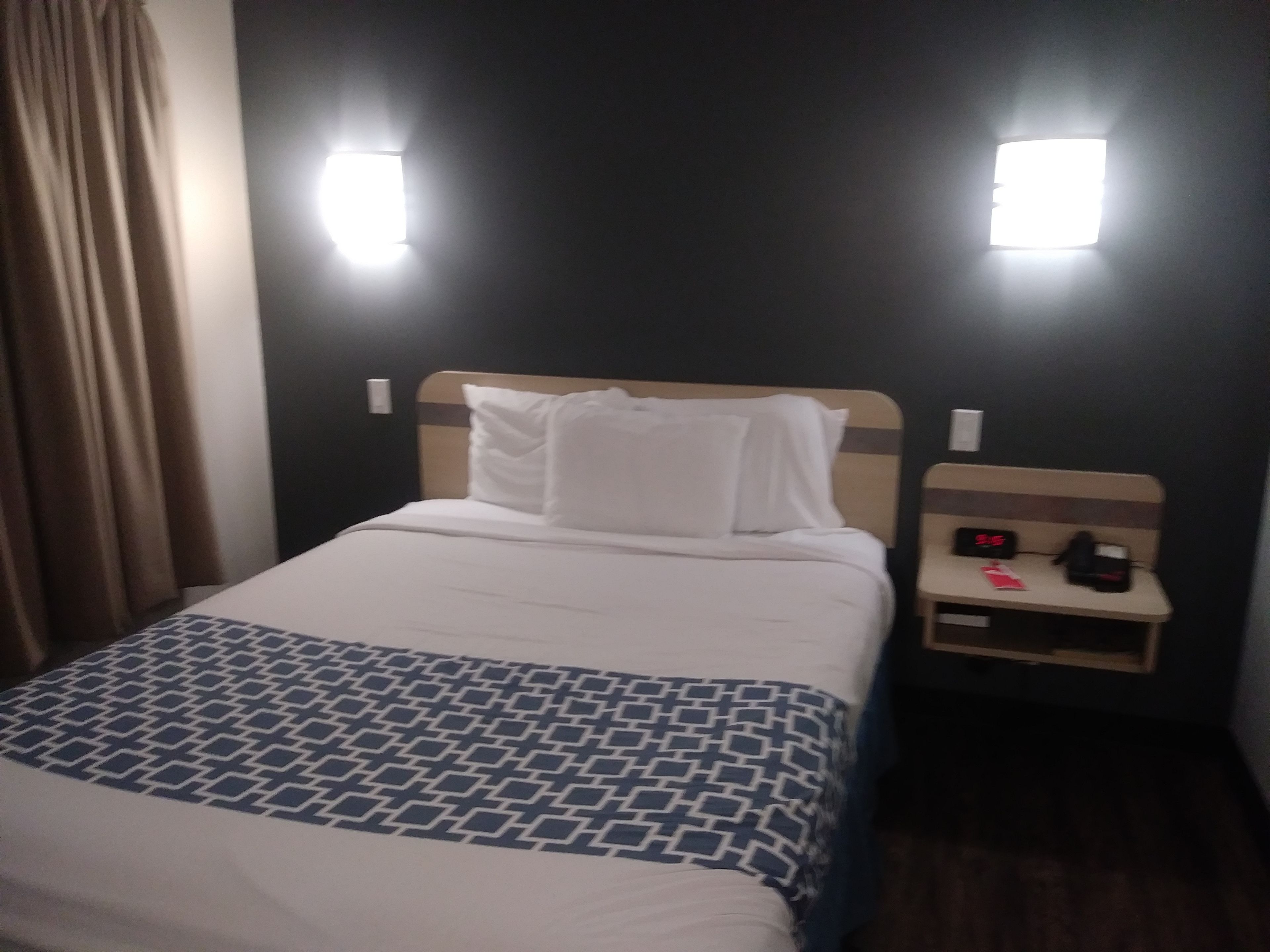 A hotel room with a bed, a nightstand, and a wall-mounted light fixture.