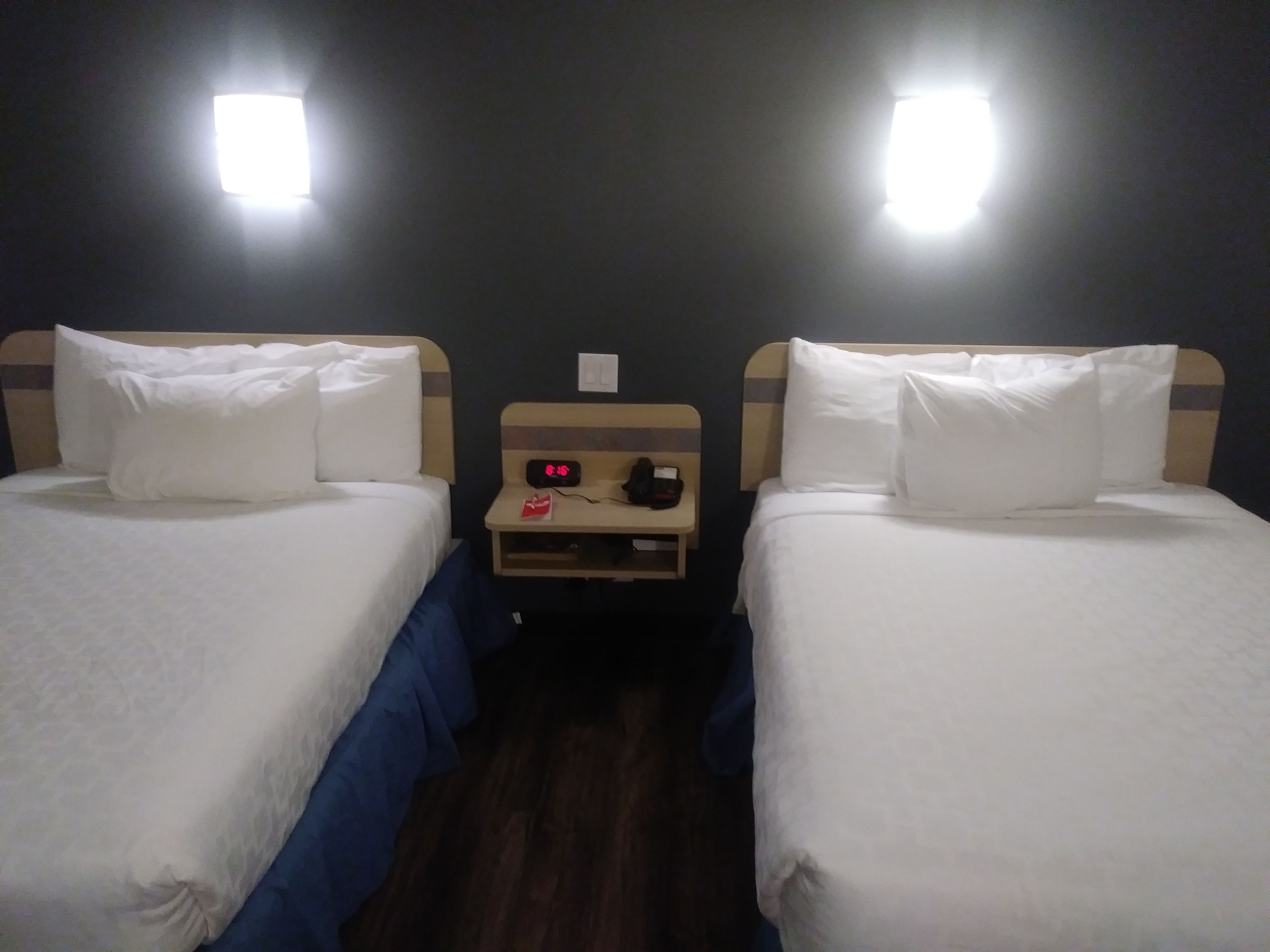 Ein Hotelzimmer mit zwei Einzelbetten, jeweils mit weißer Bettwäsche und Kissen. Dazwischen befindet sich ein Nachttisch aus Holz mit einer Uhr und einem Telefon. Der Raum hat eine dunkle Wand und zwei Wandleuchten.