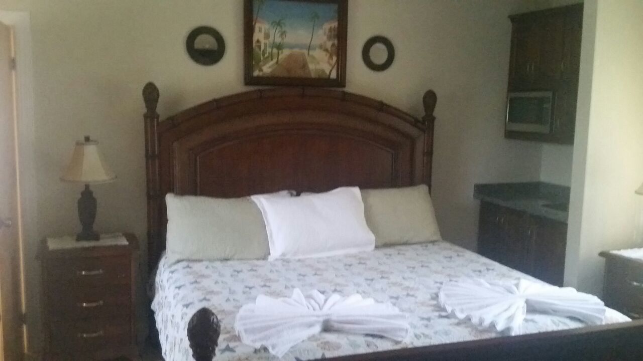 Ein ordentlich bezogenes Bett mit floralem Überwurf und zwei Kissen, ein hölzernes Kopfteil, ein Nachttisch mit Lampe und ein Gemälde an der Wand.