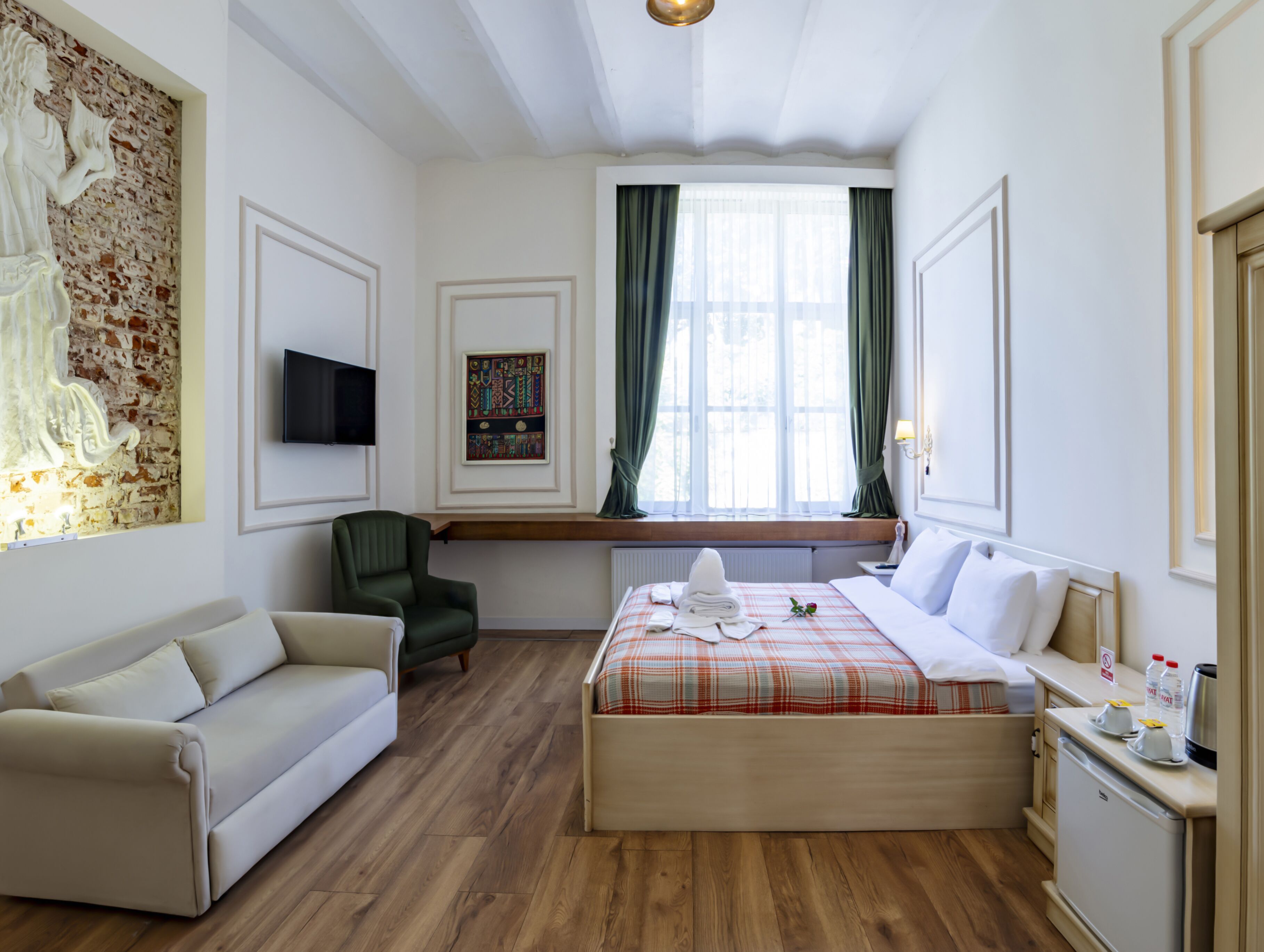A hotel room with a bed, a sofa, a chair, a TV, a window with curtains, and artwork on the walls.