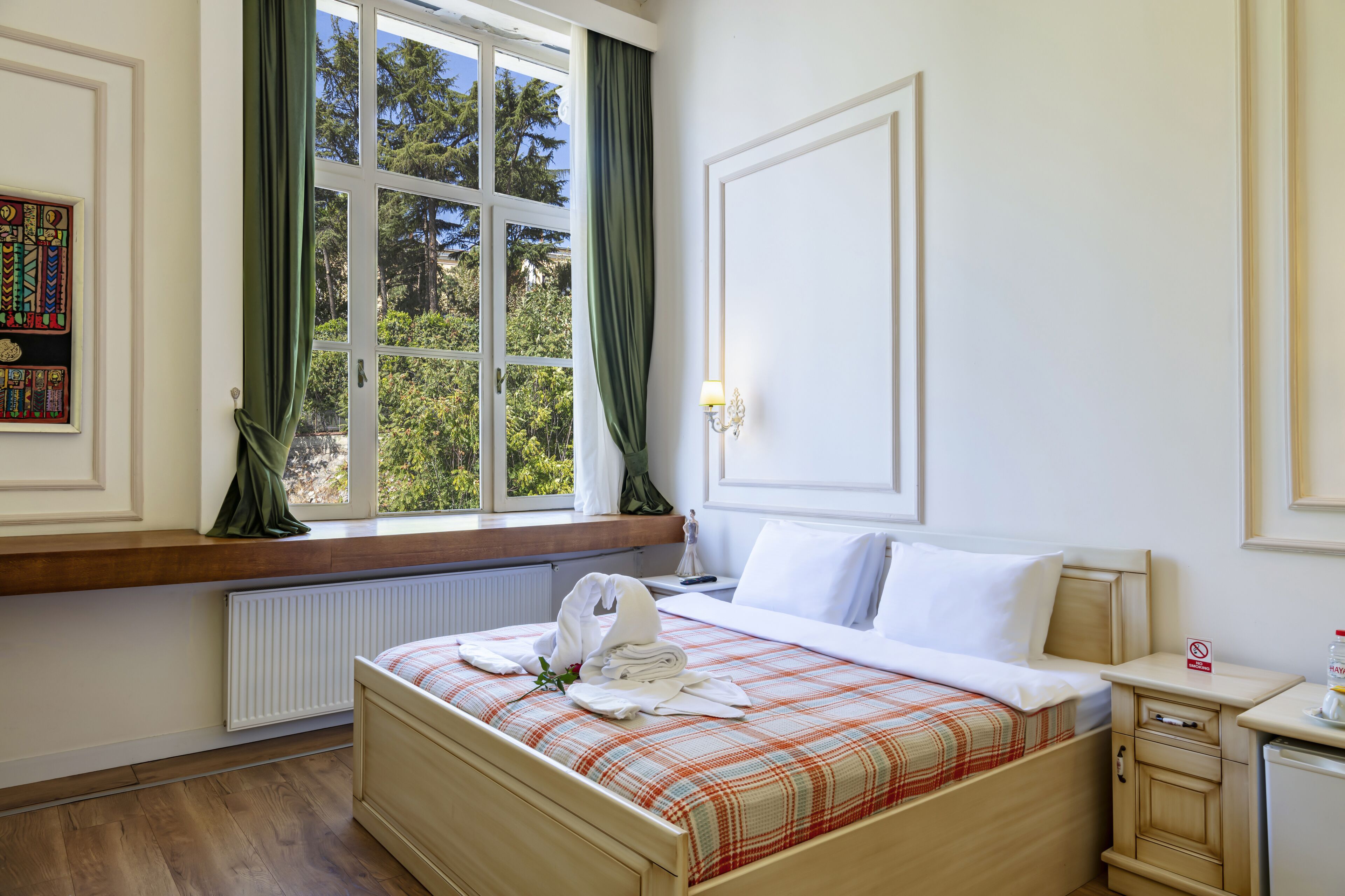 A hotel room with a bed, a window with curtains, a radiator, and a small table with a lamp.