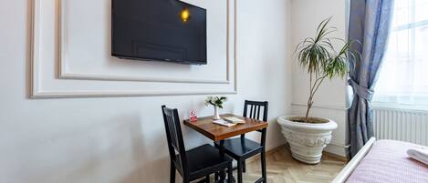Une chambre d’hôtel équipée d’un téléviseur à écran plat fixé au mur, d’une table en bois avec deux chaises, d’une plante en pot et d’un lit recouvert d’une couverture rose.