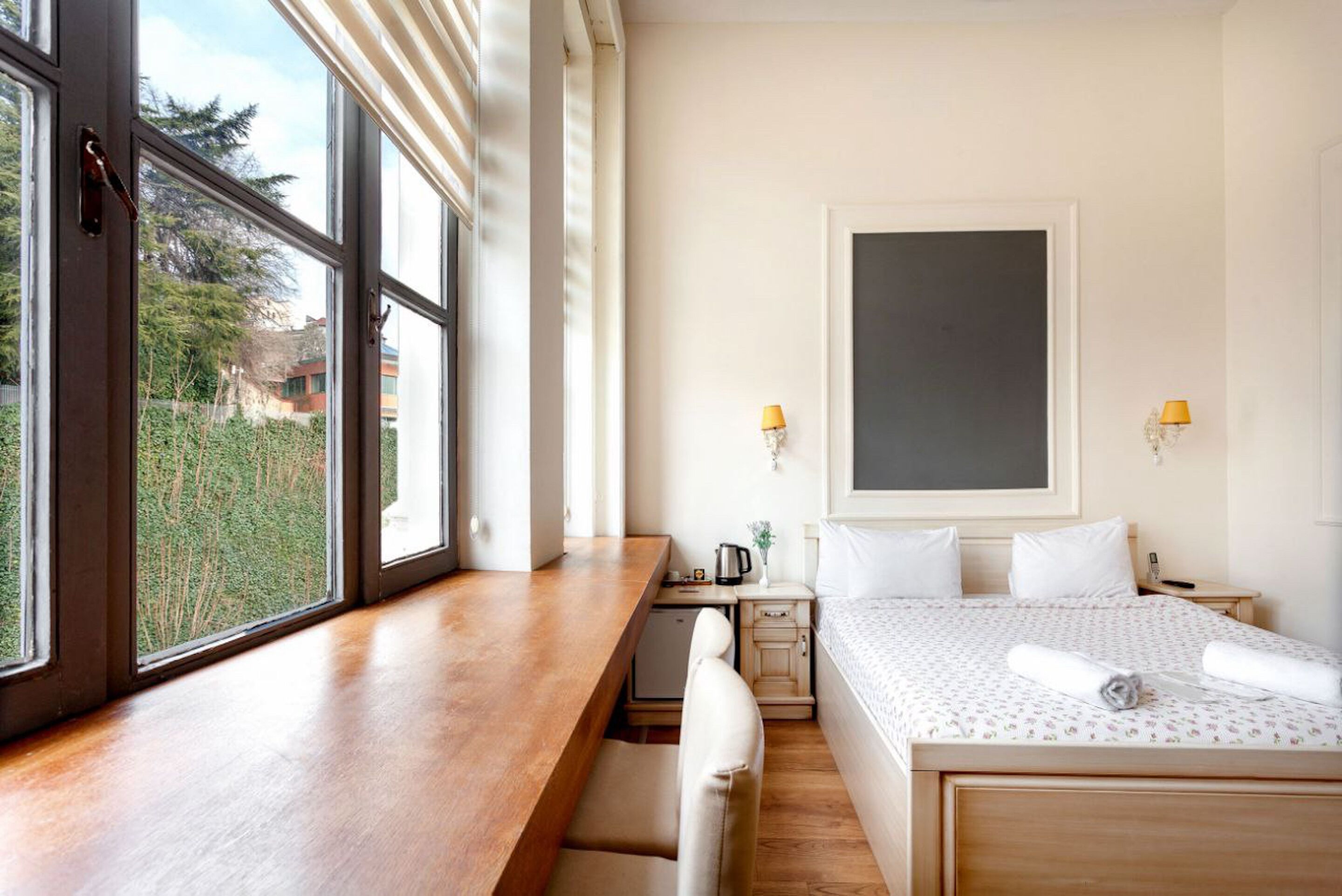 A hotel room with a wooden desk, a bed with white bedding, a nightstand, a lamp, and a view of greenery outside.