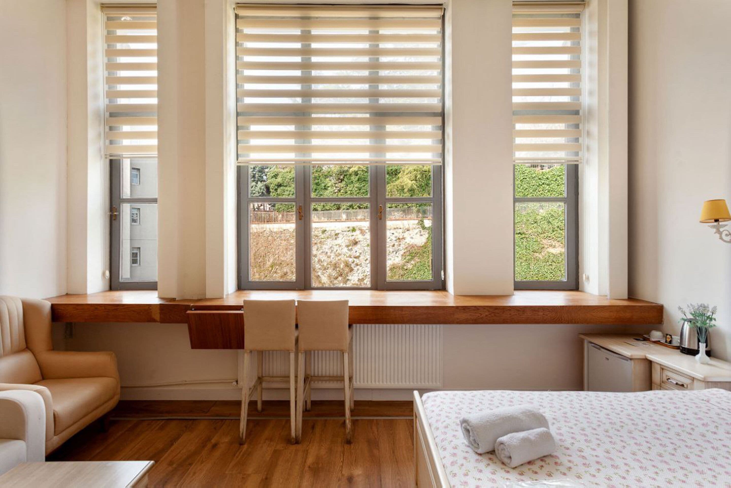 A bedroom with a wooden desk, a bed with a floral bedspread, a chair, and a view of greenery outside.