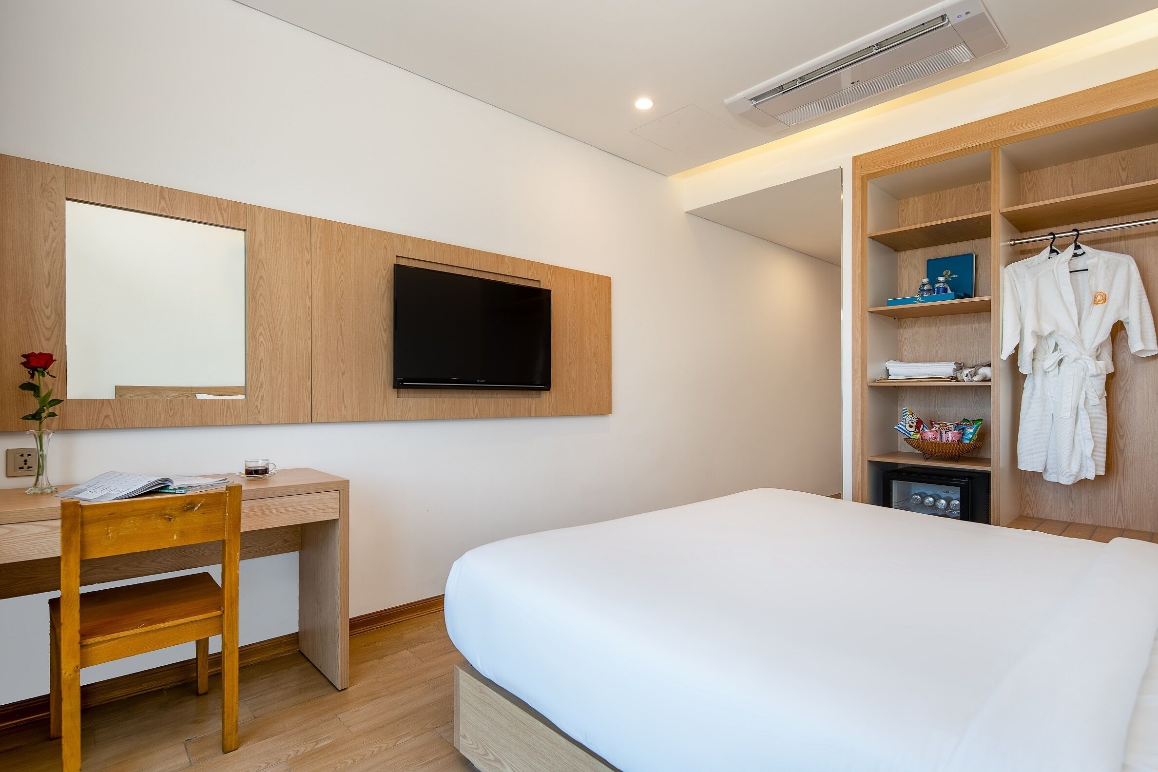 Habitación de hotel con escritorio de madera, cama, televisor montado en la pared y vestidor con bata y toallas.
