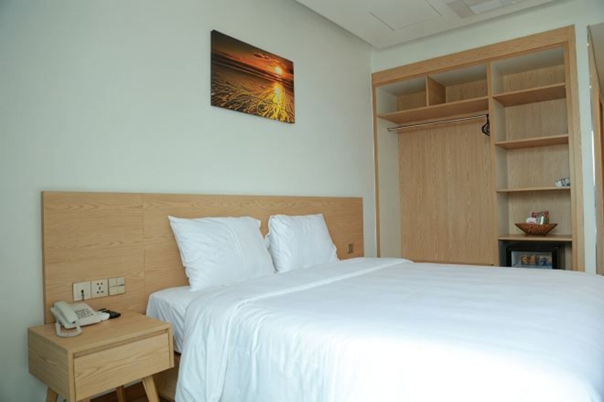 A hotel room with a bed, a nightstand, a wooden headboard, a wall-mounted artwork, and a built-in shelving unit.